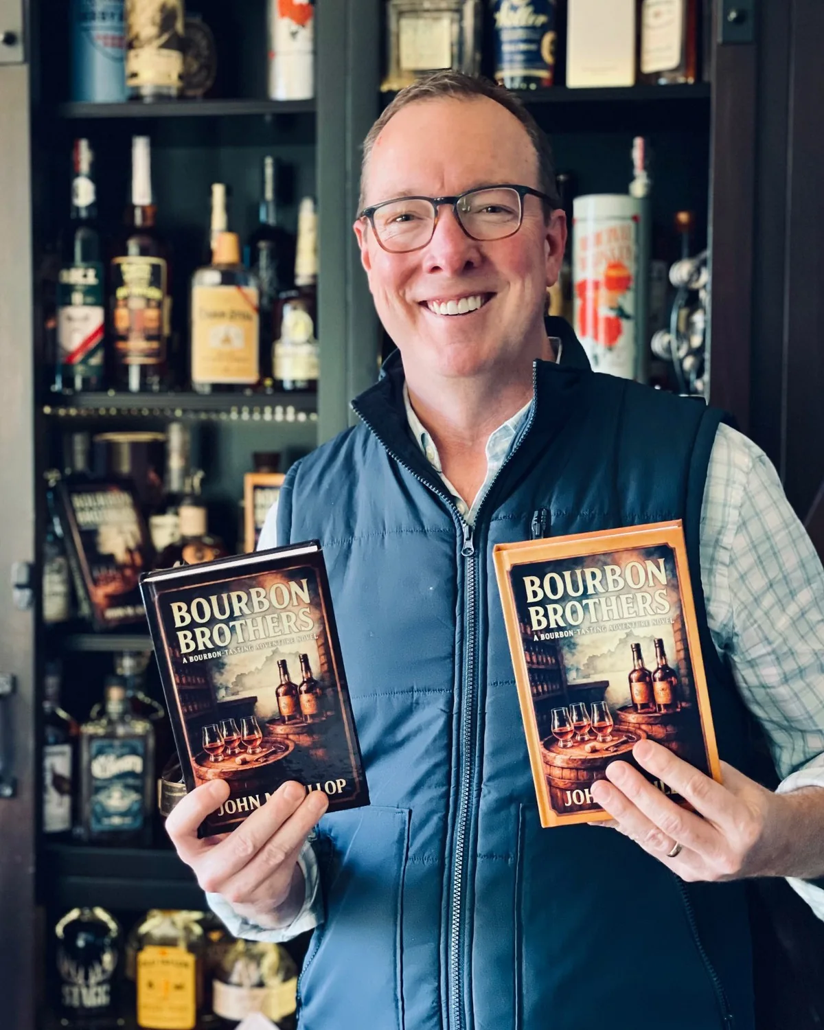 John McKillop holding two hardcover editions of Bourbon Brothers in front of his bourbon shelf
